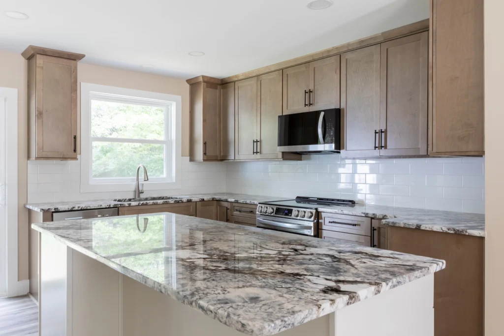 Modern kitchen with light brown cabinets, a spacious island featuring marble countertops, and a white subway tile backsplash. The room includes stainless steel appliances, a large window with a view of greenery, and hardwood flooring, creating a bright and open atmosphere.