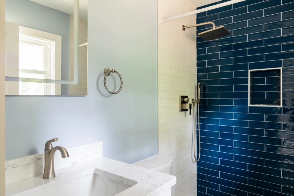 Sleek bathroom with a marble vanity sink, modern faucet, and a wall-mounted towel ring. The shower area boasts dark blue tile walls with a built-in niche, a rainfall showerhead, and a handheld sprayer, complemented by a window allowing natural light to enhance the calming ambiance.