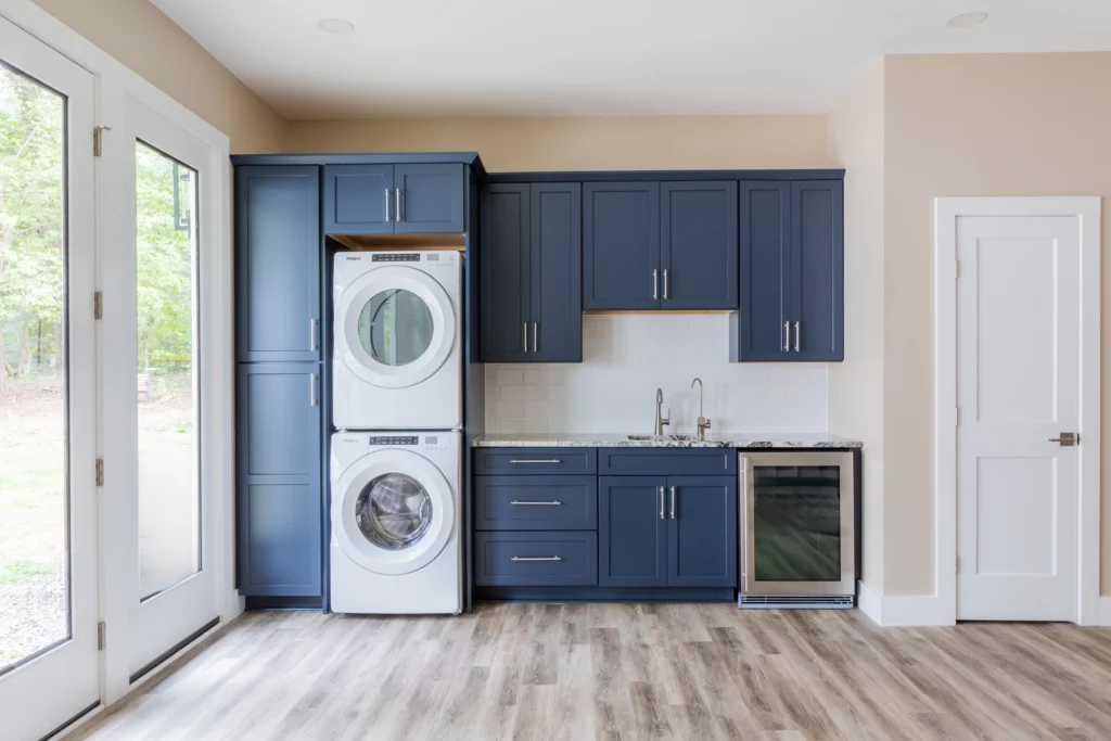 Contemporary laundry and utility area with navy blue cabinets, a stacked washer and dryer unit, and a built-in wine cooler. The space features a white subway tile backsplash, hardwood flooring, and a glass door opening to an outdoor view, blending functionality with style.
